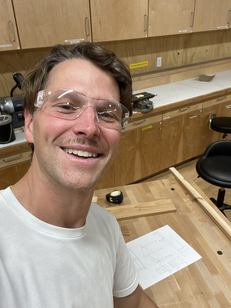 a smiling man, Ben, facing the camera with a woodworking workbench in the backround. There are tools and wooded components atop the bench.